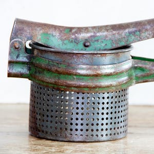 May include: An antique potato ricer with a distressed, vintage appearance. The metal tool features a perforated base, a green-painted handle, and signs of rust, suggesting age and use. A kitchen utensil for mashing potatoes.