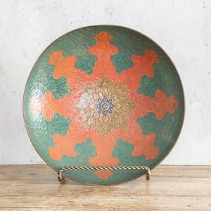 May include: A decorative, round metal plate with a green and orange floral design. The plate is supported by a metal stand and sits on a wooden surface. The plate's diameter is approximately 12 inches.