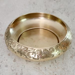 May include: A round, gold-colored bowl with floral engravings around the rim. The bowl has a smooth, reflective interior and sits on a neutral surface. The floral design adds a decorative element to the bowl.
