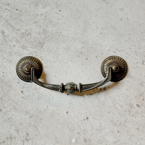 May include: A vintage-style, ornate, brass drawer pull with a rounded, decorative center piece. The pull has a smooth, slightly curved, metal arm.