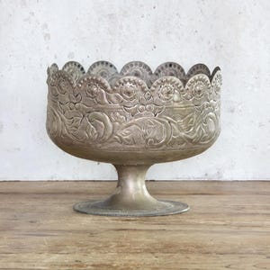 May include: Silver-colored pedestal bowl with ornate floral and scrollwork detailing. The bowl has a scalloped rim and a round base. The bowl is made of metal and sits on a wooden surface.