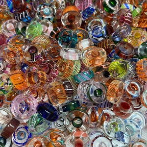 May include: A close-up shot of a collection of colorful glass beads. The beads are in various shapes and sizes, with a mix of transparent and opaque designs. Colors include orange, blue, green, pink, and brown. Some beads have striped or swirled patterns.