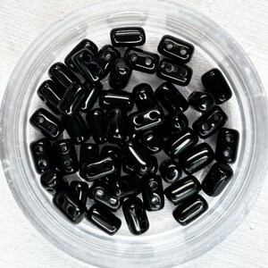 May include: A close-up of a container filled with black rectangular beads. The beads have two holes in each.
