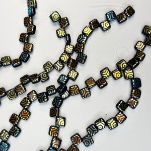 May include: A strand of black square-shaped beads with a gold geometric pattern. The beads are strung on a clear thread.