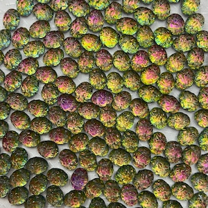 May include: A collection of iridescent, pebble-shaped beads. The beads display a range of colors, including green, yellow, purple, and pink, with a shimmering, metallic finish. The beads are scattered across a white surface, creating a textured pattern.