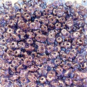 May include: Close-up of a pile of iridescent purple Czech glass beads. The beads have a unique two-hole design, and their surfaces reflect light, creating a shimmering effect. These beads are ideal for jewelry making and crafting projects.