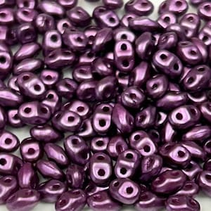 May include: A close-up view of a pile of small, purple, iridescent glass beads. The beads are shaped like teardrops with two holes at the top.