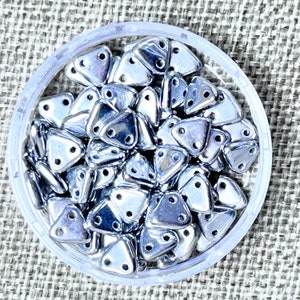 May include: A clear plastic container filled with shiny, silver-colored triangle beads. Each bead has two holes for stringing. The beads are reflective and appear to be made of metal or a metallic material. The container sits on a textured, neutral-colored surface.