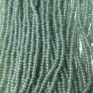 May include: Close-up of multiple strands of pale blue-green glass beads. The beads are small, round, and strung together, creating a textured, flowing appearance. The color is a soft, muted tone, suitable for jewelry making or craft projects.