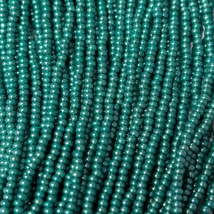 May include: Close-up of strands of teal-colored beads. The beads are small, round, and strung together, creating a textured surface. The beads have a slight sheen, reflecting light. The overall effect is a dense, shimmering display of jewelry components.