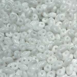 May include: A close-up view of a pile of white seed beads. The beads are small and oval-shaped with two holes.