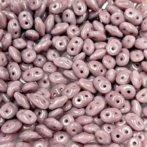 May include: A close-up view of a pile of small, light purple, glass beads. The beads are shaped like a teardrop with two holes.