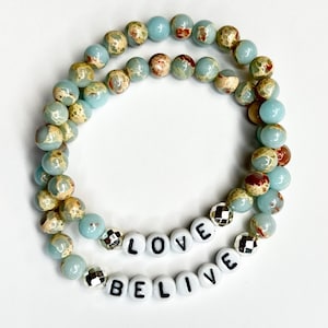 May include: A double-strand bracelet made of blue and brown patterned beads with silver spacers. The bracelet has the words "LOVE" and "BELIEVE" spelled out in white beads.