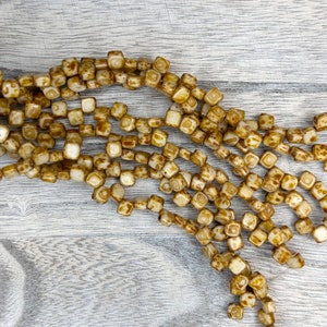 May include: A strand of square-shaped glass beads with a white base and brown speckles. The beads are strung together on a clear thread.