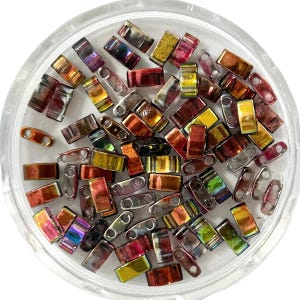 May include: A collection of colorful, rectangular beads with a metallic sheen, displayed in a clear, round container. The beads vary in color, including shades of red, gold, and iridescent hues. Each bead has two small holes, suggesting they are for stringing or crafting.