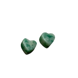 May include: Two heart-shaped jade stones with a mottled green and white pattern. The stones are smooth and polished, with a slight curve to their shape. The background is plain white.
