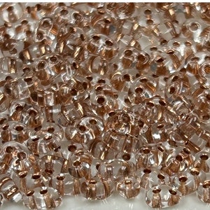 May include: A close-up of a large quantity of small, clear glass beads with a metallic gold finish. The beads have a faceted, round shape and are scattered on a white surface.