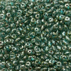 May include: A close-up shot of a pile of teal-colored beads. The beads have a metallic sheen and a unique, irregular shape with two holes. The beads are scattered, creating a textured surface. The color is a mix of teal, silver, and hints of gold.
