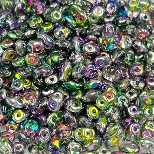 May include: A close-up view of a pile of iridescent green and purple seed beads. The beads are small and have a teardrop shape.