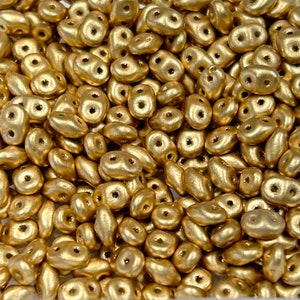 May include: Close-up of a pile of gold-colored, double-holed beads. The beads have a metallic finish and a smooth, organic shape. They are scattered, filling the frame, suggesting a craft supply or jewelry component.