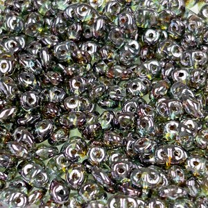 May include: A close-up of a large quantity of small, iridescent green glass beads. The beads are shaped like teardrops with a hole in the center. They are arranged in a random pattern.