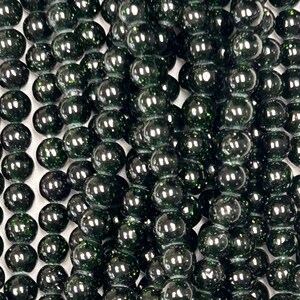 May include: Close-up of a collection of dark green, glossy beads. The beads are round and appear to be strung together, creating a textured surface. The beads have a reflective quality, with highlights of light across their surfaces.