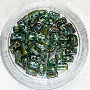 May include: A close-up of a container filled with green and brown glass beads. The beads are rectangular with two holes in each. The beads are iridescent and have a speckled appearance.