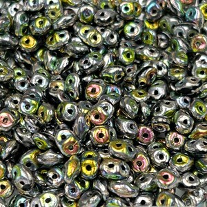 May include: A close-up of a pile of small, silver-colored glass beads with iridescent green and purple accents. The beads are shaped like teardrops with two holes.