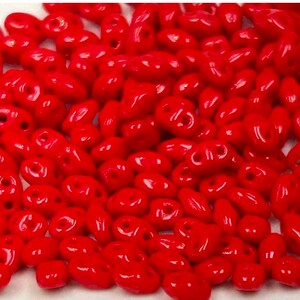 May include: A large pile of bright red seed beads. The beads are small and oval shaped with two holes in each bead.
