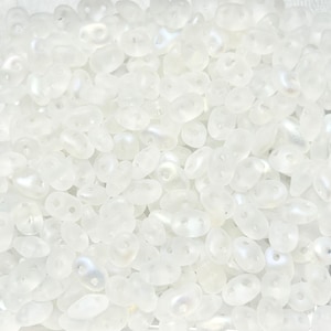 May include: A close-up view of a large quantity of small, white, opaque, glass beads. The beads are shaped like a teardrop with two holes at the top.