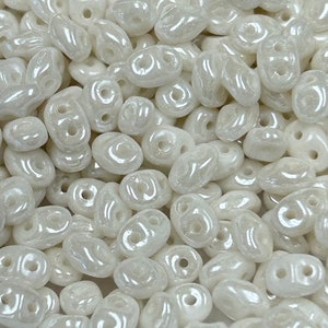 May include: A close-up of a pile of white, opaque, oval-shaped beads with two holes each. The beads have a slightly iridescent finish.