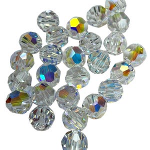 May include: A collection of clear, faceted crystal beads. The beads have an iridescent coating that reflects a rainbow of colors, including yellow, blue, and purple. The beads are round with multiple facets, creating a sparkling effect.