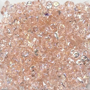 May include: A close-up of a pile of iridescent pink glass beads. The beads are small and round, with a slightly irregular shape. They have a subtle shimmer and sparkle.