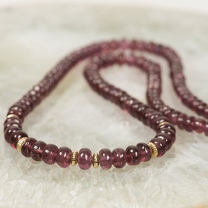 Puede incluir: Un collar largo hecho de peque&ntilde;as cuentas redondas de color p&uacute;rpura con detalles dorados. El collar est&aacute; sobre una superficie blanca texturizada.