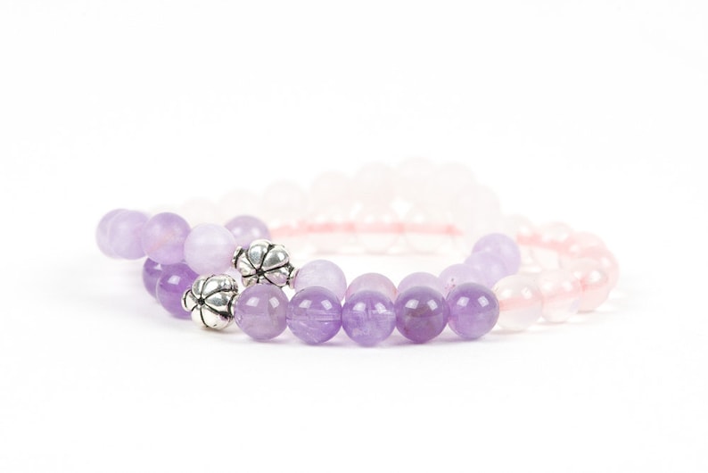 May include: Two bracelets made of amethyst and rose quartz beads. The amethyst bracelet has silver flower-shaped beads.