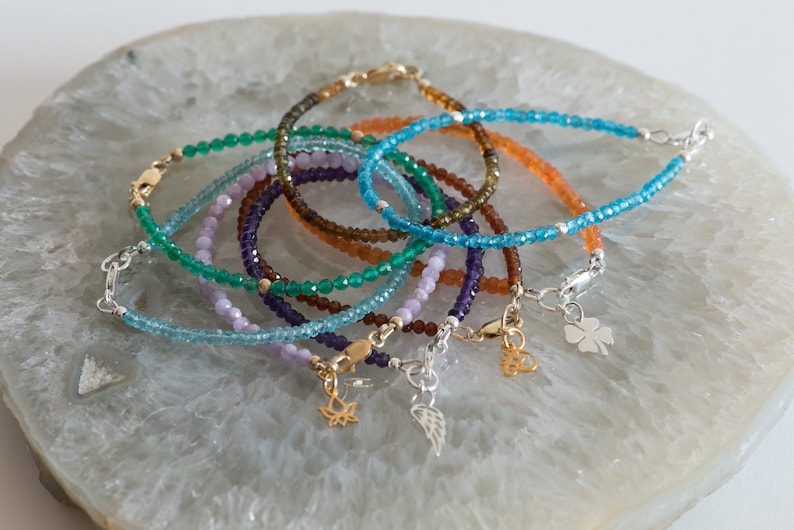 Peut inclure: Un ensemble de sept bracelets en pierres pr&eacute;cieuses avec des breloques en argent et en or. Les bracelets sont faits de pierres pr&eacute;cieuses de diff&eacute;rentes couleurs, notamment le vert, le bleu, le violet et le brun. Les breloques comprennent une plume, une fleur de lotus et un tr&egrave;fle &agrave; quatre feuilles.