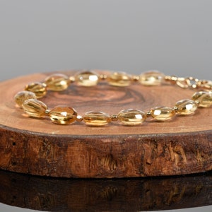 May include: A gold-toned bracelet made of oval-shaped beads. The bracelet is on a wooden surface.