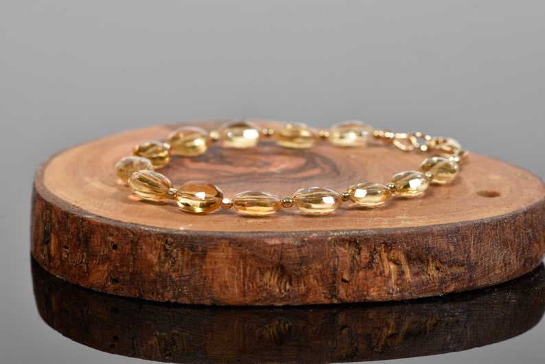May include: A gold-toned bracelet made of oval-shaped beads. The bracelet is on a wooden surface.