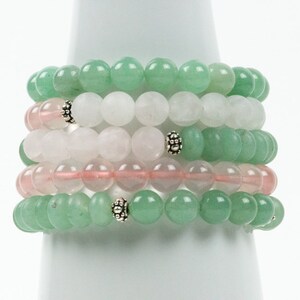 Op de afbeelding: Stapel van zes armbanden gemaakt met groene, witte en roze kralen. De armbanden zijn gestapeld op een witte displaystandaard.