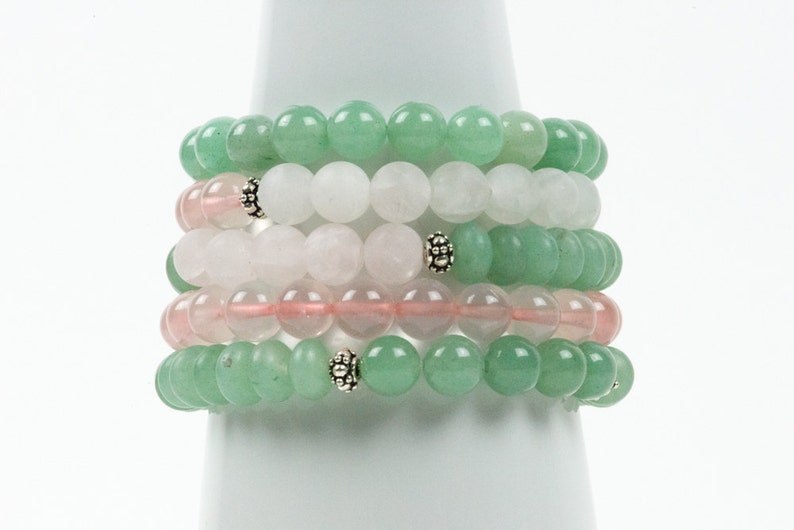 Op de afbeelding: Stapel van zes armbanden gemaakt met groene, witte en roze kralen. De armbanden zijn gestapeld op een witte displaystandaard.