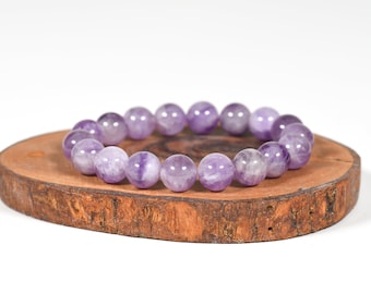 Puede incluir: Un brazalete hecho de cuentas redondas de amatista pulida. Las cuentas son de color púrpura claro con vetas blancas. El brazalete descansa sobre una pequeña rodaja de madera redonda con borde natural. El fondo es blanco.