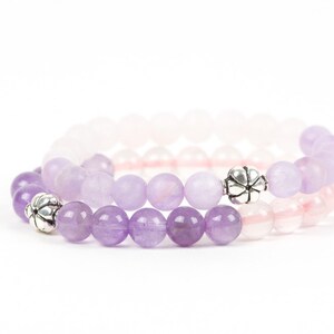 May include: Two bracelets made of amethyst and rose quartz beads with silver flower-shaped charms. The amethyst beads are a darker purple and the rose quartz beads are a lighter pink.