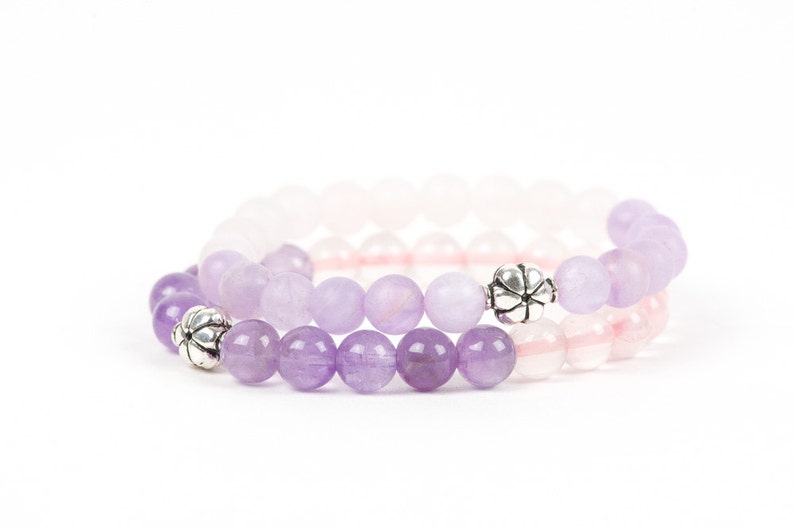 May include: Two bracelets made of amethyst and rose quartz beads with silver flower-shaped charms. The amethyst beads are a darker purple and the rose quartz beads are a lighter pink.