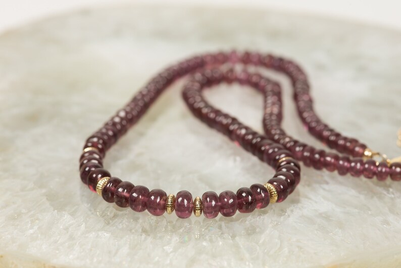 Puede incluir: Un collar largo hecho de peque&ntilde;as cuentas redondas de color p&uacute;rpura con detalles dorados. El collar est&aacute; sobre una superficie blanca texturizada.