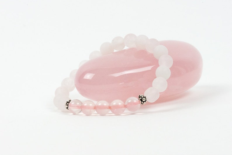 Op de afbeelding: Een armband van witte en roze edelstenen met zilveren kralen. De armband ligt op een roze rozenkwarts steen.