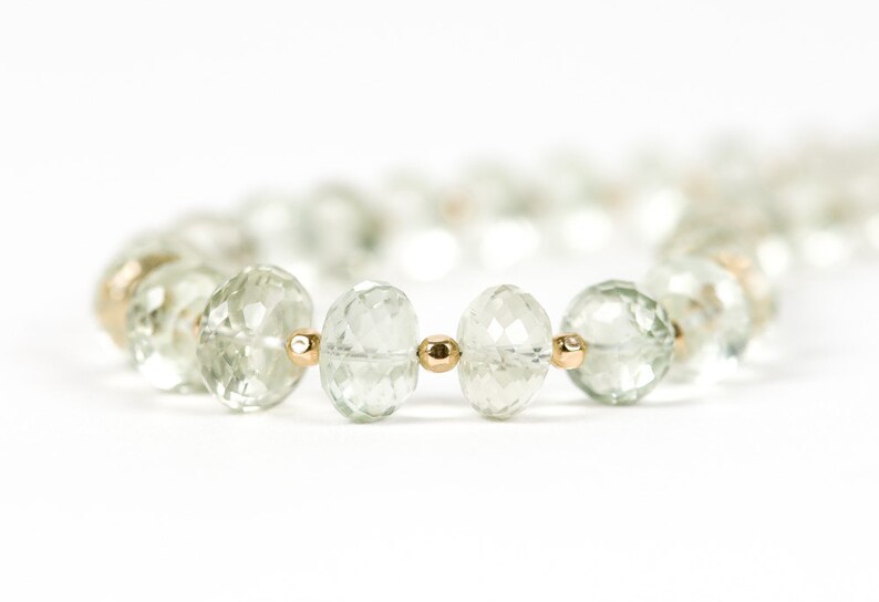 May include: A close-up of a green gemstone necklace with gold beads. The necklace is made of faceted, round gemstones and is strung on a gold chain.