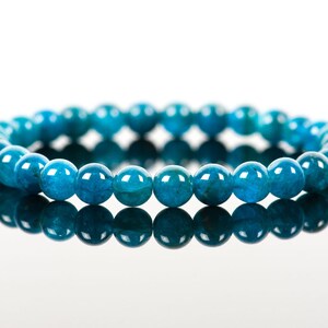 May include: A blue gemstone bracelet made of round beads. The bracelet is lying on a white surface and is reflecting in the surface.