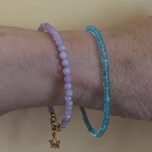 Peut inclure: Deux bracelets en perles, l'un violet clair et l'autre bleu clair, port&eacute;s au poignet. Le bracelet violet a un charme en or en forme de fleur de lotus.