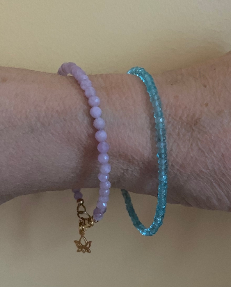 Peut inclure: Deux bracelets en perles, l'un violet clair et l'autre bleu clair, port&eacute;s au poignet. Le bracelet violet a un charme en or en forme de fleur de lotus.