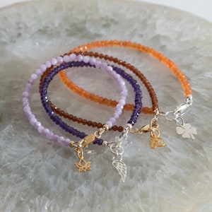 Peut inclure: Trois bracelets en perles avec des breloques. Les bracelets sont superpos&eacute;s. Le bracelet du haut est orange, le bracelet du milieu est violet et le bracelet du bas est violet clair. Les breloques sont une fleur de lotus dor&eacute;e, une aile argent&eacute;e et un tr&egrave;fle &agrave; quatre feuilles argent&eacute;.